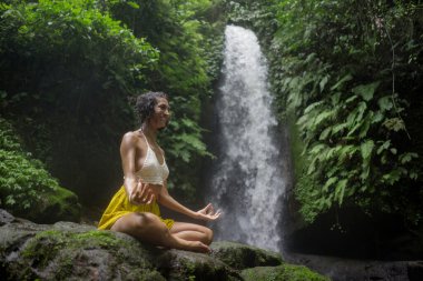Genç çekici ve mutlu hippi kadın dışarıda yoga yapıyor tropikal şelalede meditasyon yapıyor sağlıklı ve zen yaşam tarzında özgürlük ve doğanın tadını çıkarıyor.