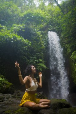 Genç çekici ve mutlu hippi kadın dışarıda yoga yapıyor tropikal şelalede meditasyon yapıyor sağlıklı ve zen yaşam tarzında özgürlük ve doğanın tadını çıkarıyor.