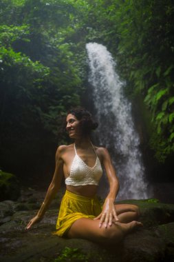 Genç çekici ve mutlu hippi kadın dışarıda yoga yapıyor tropikal şelalede meditasyon yapıyor sağlıklı ve zen yaşam tarzında özgürlük ve doğanın tadını çıkarıyor.