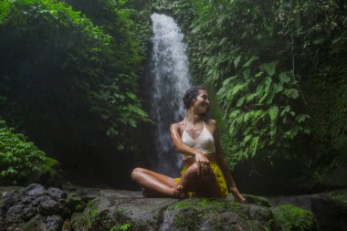 Genç çekici ve mutlu hippi kadın dışarıda yoga yapıyor tropikal şelalede meditasyon yapıyor sağlıklı ve zen yaşam tarzında özgürlük ve doğanın tadını çıkarıyor.
