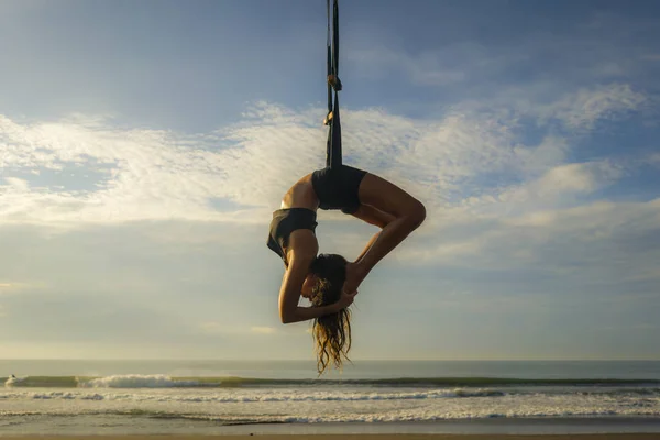 Aero yoga beach workout - silhouette of young attractive and athletic woman practicing aerial yoga exercise training acrobatic  body postures on blue sky over sea — Stock Photo, Image
