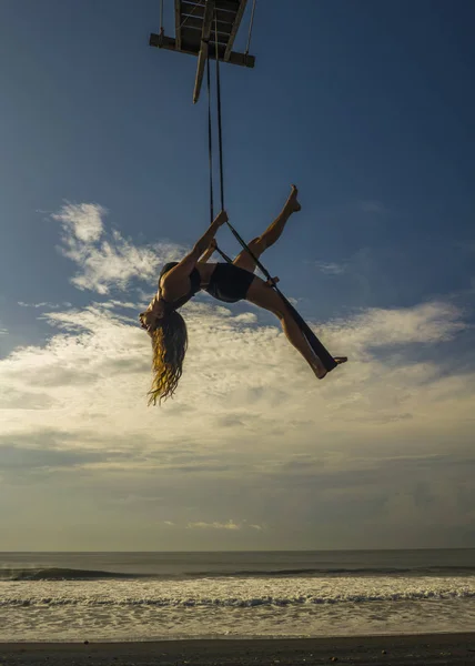 Aero ioga praia treino - silhueta de jovem atraente e atlética mulher praticando exercícios de ioga aérea treinamento acrobático posturas do corpo no céu azul sobre o mar — Fotografia de Stock