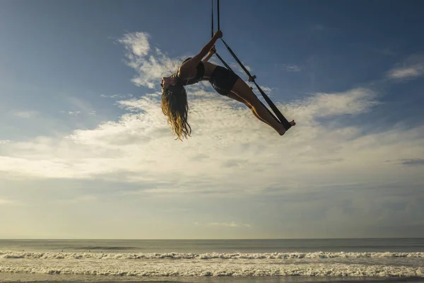 Entrenamiento de playa de yoga aéreo - silueta de mujer joven atractiva y atlética que practica yoga aéreo entrenamiento de ejercicios posturas corporales acrobáticas en el cielo azul sobre el mar — Foto de Stock