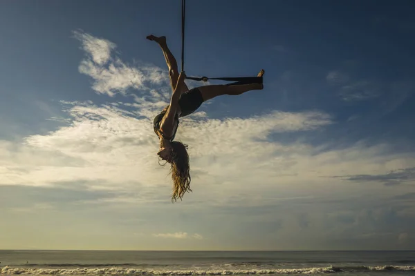 Entrenamiento de playa de yoga aéreo - silueta de mujer joven atractiva y atlética que practica yoga aéreo entrenamiento de ejercicios posturas corporales acrobáticas en el cielo azul sobre el mar — Foto de Stock