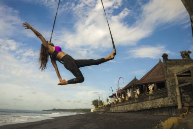 Aero yoga plaj egzersizi - Bali adasında deniz üzerinde mavi gökyüzünde akrobatik vücut hareketleri eğitimi alan çekici ve atletik kadın 