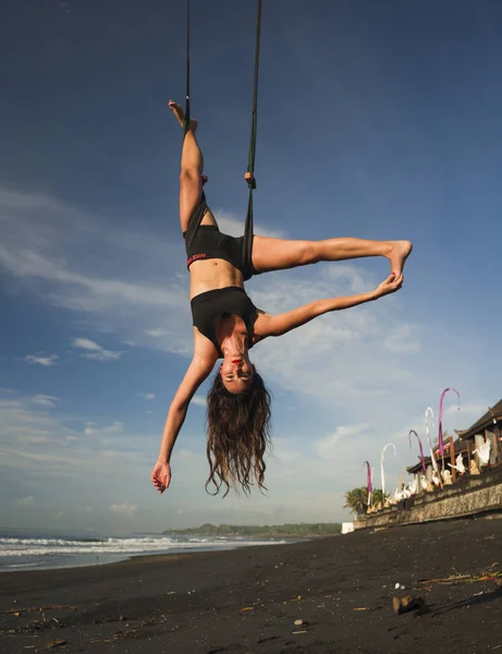 Aero yoga beach workout - giovane donna attraente e atletica che pratica esercizi di yoga aereo formazione acrobatica posture del corpo sul cielo blu sul mare a Bali Island — Foto Stock