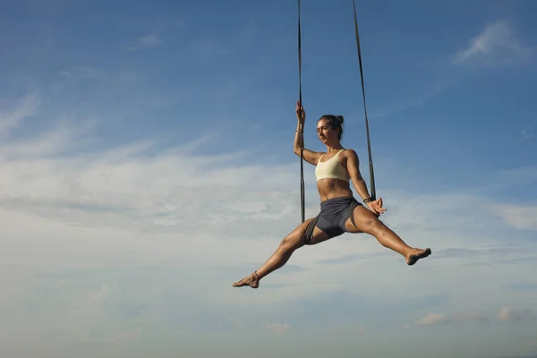 Aerial yoga meditation workout  isolated on blue sky - young attractive and healthy woman practicing aero-yoga training balance body and mind control hanging from ropes — Stock Photo, Image