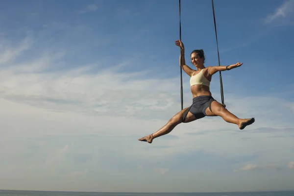 Aerial yoga beach workout - young attractive and healthy woman practicing aero-yoga training balance body and mind control hanging from ropes above the sea — Stock Photo, Image