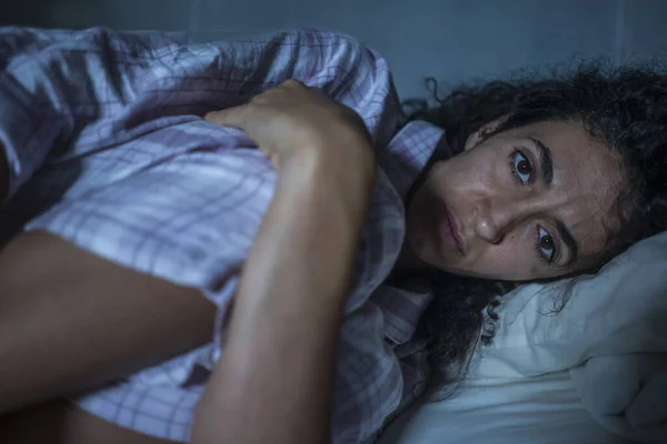 Young sad and depressed hispanic woman with curly hair sleepless in bed suffering excruciating period pain holding her belly in fetal position — Stock Photo, Image