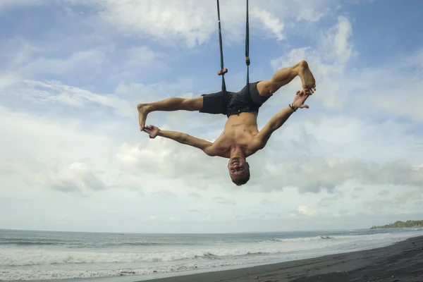 Ao ar livre retrato de jovem atraente e atlético homem praticando aero ioga workoutat praia pendurado de corda swiing acima do mar formação equilíbrio corporal e flexibilidade — Fotografia de Stock