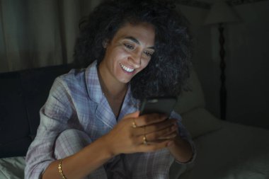 Genç, mutlu ve çekici hipster kadın geceleri yatakta rahat cep telefonuyla internet üzerinden sosyal medya uygulamasının keyfini çıkarıyor ve hücre bağımlılığı konseptinde neşeli gülümsüyor.