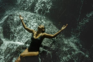 Genç, çekici ve mutlu Asyalı Endonezyalı kadın tropikal ormanda doğal şelalenin güzelliği karşısında mutlu ve egzotik yolculuğun tadını çıkarıyor.