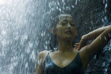 Genç, çekici ve mutlu Asyalı Endonezyalı kadın tropikal ormanda doğal şelalenin güzelliği ve saflığı karşısında mutlu ve egzotik bir yolculuğun tadını çıkarıyor.