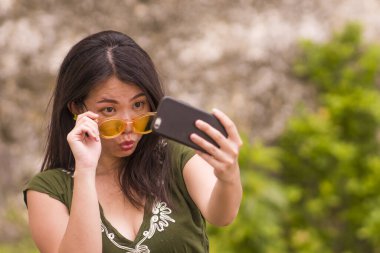 Genç ve güzel Asyalı kadın cep telefonuyla selfie çekiyor. Hipster güneş gözlükleriyle neşeli ve tasasız poz veriyor. Yaz tatilinin tadını çıkarıyor.