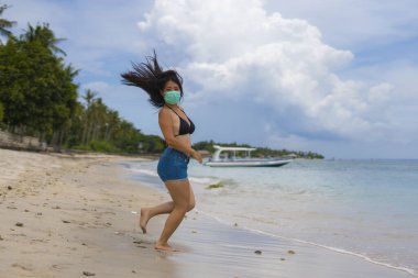 Halka açık yerlerde tıbbi maske kullanmak. Genç, çekici, Asyalı bir kadın. Plaj tatillerinde bikini giyiyor ve virüs enfeksiyonuna karşı koruyucu maske takıyor. 