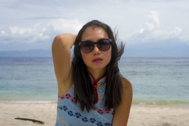 Güzel ve mutlu Asyalı Çinli kadın şık yaz elbiseli ve güneş gözlüklü. Tropik plajda turist gibi rahatlamış seyahat ve tatil konseptinde dinleniyor.