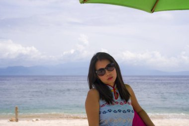 Şık yaz elbiseli ve güneş gözlüklü genç, güzel ve mutlu Asyalı kadın tropik plajda rahatlamış bir şekilde şemsiye altında puf çantanın üzerinde rahatça oturuyordu.