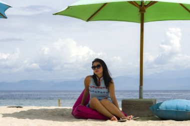 Şık yaz elbiseli ve güneş gözlüklü genç, güzel ve mutlu Asyalı kadın tropik plajda rahatlamış bir şekilde şemsiye altında puf çantanın üzerinde rahatça oturuyordu.