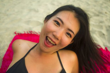 Açık havada, güzel ve mutlu Asyalı bikinili genç bir kadının plaj kumsalındaki sargısına uzanmış neşeli ve rahat bir şekilde güzellik ve moda kavramında gülümseyen doğal portresi.