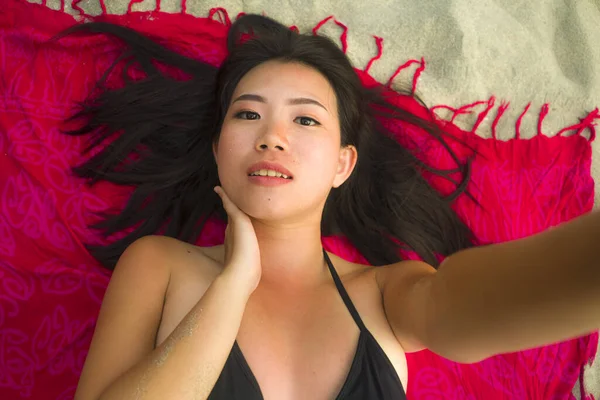 outdoors fresh and natural portrait of young beautiful and happy Asian Korean woman in bikini lying flat on sarong on beach sand taking selfie photo with mobile smiling cheerful and relaxed