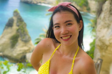 Güzel ve mutlu Asyalı bikinili bir kadın yaz tatili boyunca tropik cennet kumsalında elmas şekilli kaya uçurumuyla deniz manzarasının keyfini çıkarıyor.