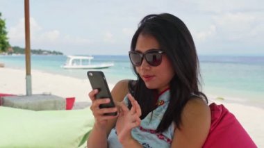Güzel ve mutlu bir Asyalı kadının üzerinde ağır çekim. Plaj otelinde puf puf çantasına oturmuş cep telefonuyla selfie çekiyor ve yaz tatilinin tadını çıkarıyor.