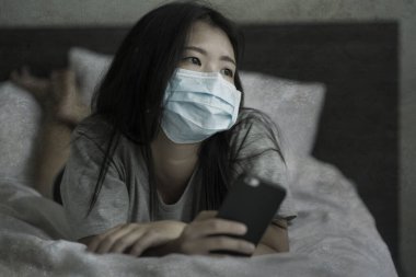 Salgın ve virüs salgını. Genç, korkmuş, endişeli, tıbbi maskeli Asyalı kadın ev hapsi ve karantina sırasında cep telefonuyla internet haberlerini kontrol ediyor.