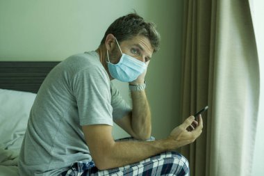 virus outbreak - dramatic portrait of sick man in medical facial mask sitting on bed scared and worried infected by coronavirus suffering lockdown alone at home in quarantine clipart