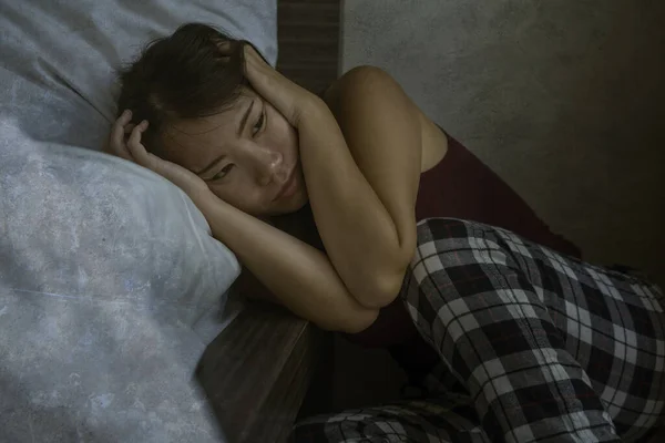dramatic dark portrait of young attractive depressed and sad Asian Korean woman on bedroom floor feeling worried and scared suffering depression problem and anxiety leaning on bed