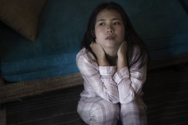 Evinde pijamalarıyla oturan üzgün ve üzgün Asyalı genç bir kadının dramatik karanlık portresi. Oturma odasındaki kanepede oturuyor. Mutsuz ve kafası karışık, kırık kalp ağrısı çekiyor.