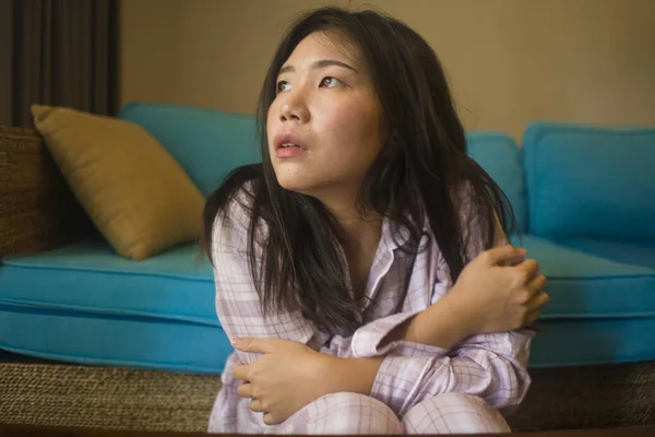 indoors lifestyle portrait of young beautiful sad and depressed Asian Korean woman in pajamas at home sitting by living room couch feeling unhappy and confused suffering broken heart pain