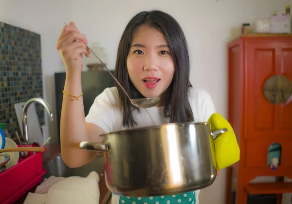 Beautiful domestic chef at work - lifestyle portrait of young pretty and happy Asian Chinese woman in apron holding cooking pot tasting soup smiling satisfied as home cook housewife