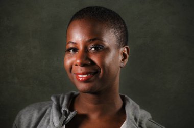 Kapüşonlu kazaklı, saçları tıraşlı, mutlu ve kendine güvenen genç, güzel, Afro-Amerikan kadın koyu arka planda izole edilmiş bir şekilde pozitif bir gülümsemeyle kameraya bakıyor.