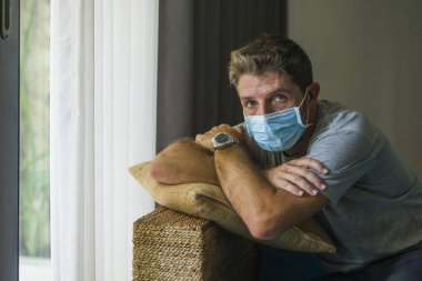 Covid-19 virüs tecridi - 30 'lu ya da 40' lı yaşlarda üzgün ve endişeli bir adam tıbbi maske takmış, evde kalıp virüs salgınını kontrol altına alma talimatlarını takiben karantinada düşünüyor ve korkuyor.