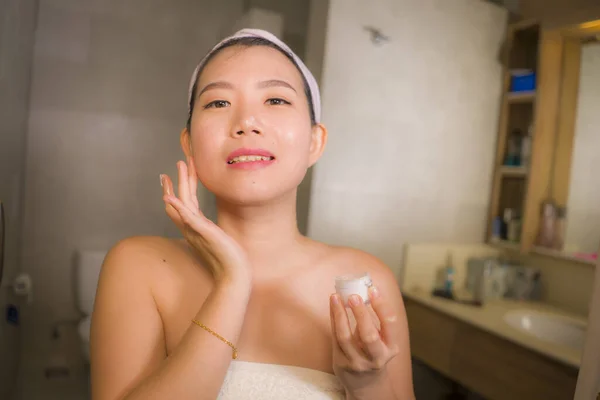 home lifestyle portrait of young happy and beautiful Asian Korean woman at bathroom mirror applying skin care moisturizer and anti-aging product in beauty and skincare concept