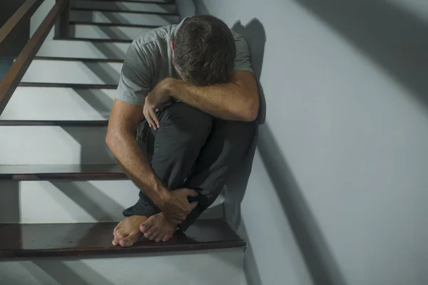 horror movie style portrait of sad and desperate man suffering depression problem or mental disorder sitting on staircase at home hopeless crying overwhelmed and helpless
