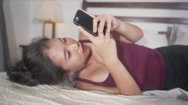 Genç, güzel ve mutlu Asyalı kadın cep telefonu sosyal medya kullanıyor erkek arkadaşıyla mesajlaşıyor ya da online flört uygulamasının keyfini çıkarıyor evde yatarken neşeyle gülümsüyor. 
