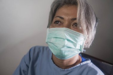 Covid-19 virüsü hastalık yayıyor. Koronavirüs enfeksiyonu kapmış, hasta önlüklü ve maskeli, korkmuş ve üzgün olgun bir kadın. İyileşme umudu içinde.