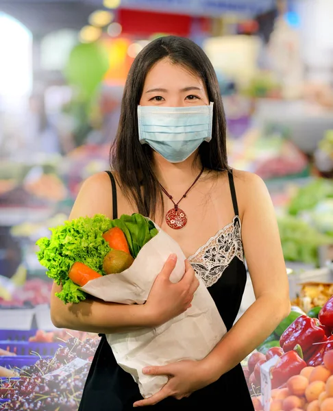 food and groceries shooping during covid-19 virus quarantine - young beautiful and positive Asian Chinese woman in face mask carrying vegetables bag stock up during coronavirus lockdown