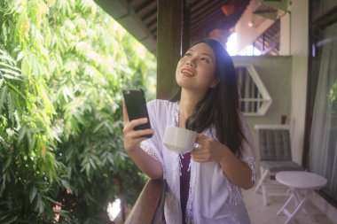 Açık havada güzel ve mutlu Asyalı bir kadının sabah kahvesini içerken çekilmiş bir portresi. Ev terasında ya da otel odasının balkonunda cep telefonunda sosyal medya kullanıyor.