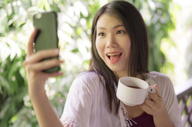 Açık havada genç, güzel ve mutlu Asyalı bir kadının portresi sabah kahvesi içerken cep telefonuyla ev terasında ya da otel odasının balkonunda selfie çekiyor.