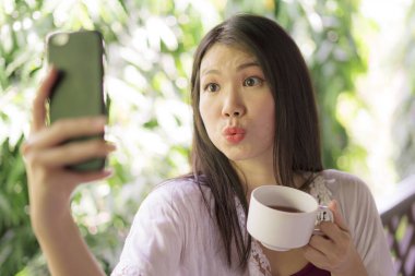 Açık havada, güzel ve mutlu Asyalı bir kadının sabah kahvesi içerken, sosyal medya için cep telefonuyla ev terasında ya da otel odasının balkonunda selfie çekerken çekilmiş resmi.