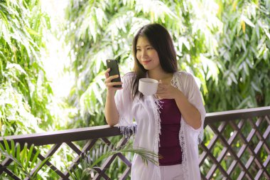 Güzel ve mutlu Asyalı bir kadının açık hava yaşam tarzı portresi. Ev terasında ya da otel odasının balkonunda internet sosyal medya kullanarak sabah kahvesi içiyor.