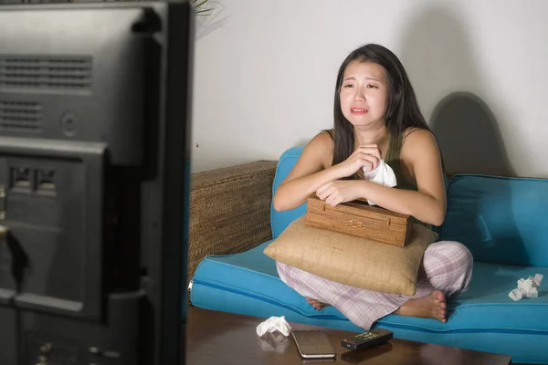 funny lifestyle portrait of young cute and emotional Asian Chinese woman watching korean drama show on TV sitting at home living room couch crying wiping tears with tissue paper