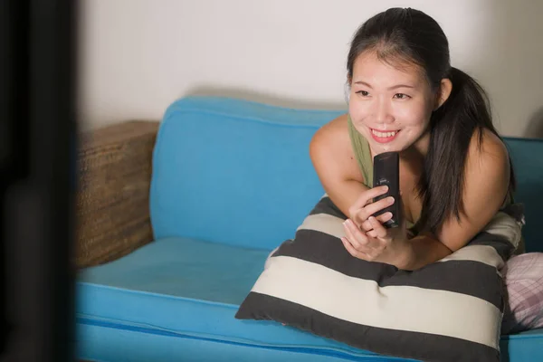 home lifestyle portrait of young happy and beautiful Asian Chinese woman watching TV  at living room sofa couch enjoying funny television Korean drama show or comedy movie relaxed and cozy