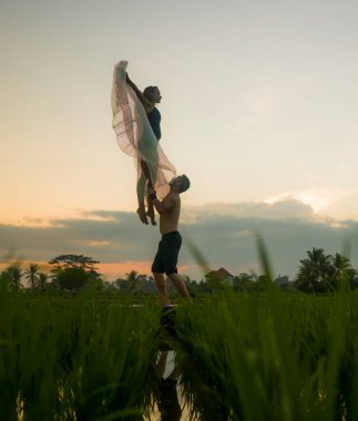 Açık havada gün batımı akroyogası egzersizi. Genç mutlu ve zinde çift, güzel pirinç tarlasında akro yoga talimi yapıyor. Doğanın tadını çıkarıyor, özgürlük kavramında kanatlı bir kadınla akrobatik poz veriyor.