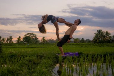 Açık havada günbatımı akroyogası silueti. Genç mutlu ve zinde çift güzel pirinç tarlasında akro yoga talimi yapıyor. Doğanın ve sağlıklı yaşam tarzının tadını çıkarıyor. 