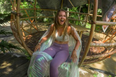 Açık havada genç güzel ve mutlu bir kızın doğal portresi. Yoga çalışmasından sonra rahatlamış. Çardakta oturmuş, tropikal ormanda neşeyle gülümsüyor.