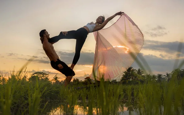 outdoors sunset acroyoga workout - young happy and fit couple practicing acro yoga drill at beautiful rice field enjoying nature and healthy lifestyle doing acrobatic pose