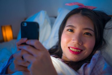 Saçında bantla ve pijamalarıyla genç, güzel ve mutlu Asyalı Çinli kız geceleri cep telefonuyla internet bağımlılığında sosyal medyada oyun oynuyor.